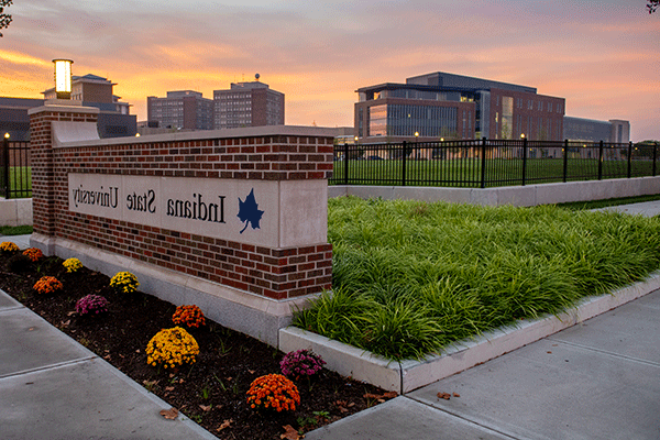 A brick stone sign reading “Indiana State University” with green grass and colorful flowers surrounding it. In the distance is the College of Health and Human Services building, a brick building with many glass windows. Other buildings are in the background.